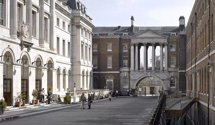 A view of the Strand Campus of King’s College London, featuring a historic courtyard flanked by classical and Georgian-style buildings. The left side has a white neoclassical façade with arched windows and decorative statues, while the right side consists of a red-brick building with a traditional design. In the background, a grand structure with large columns and an arched passageway frames the entrance to another section of the campus. Several people are walking along the paved courtyard, which is lined with potted plants and benches.